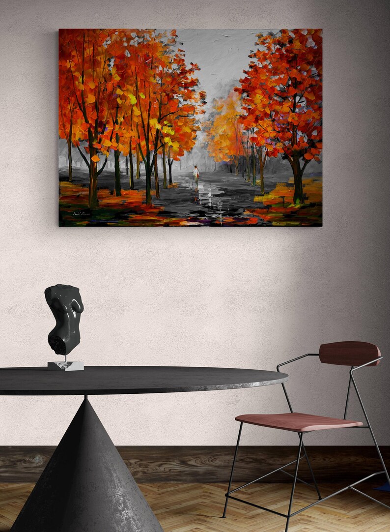 May include: An oil painting depicts a path lined with trees in autumn. The trees have vibrant orange and yellow leaves. A figure walks down the path. The painting hangs above a black table with a sculpture and a chair.