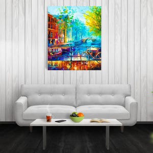 May include: An abstract painting of a European city scene with a bridge, buildings, and a bicycle. The artwork features vibrant colors, including blues, oranges, and yellows, creating a lively and artistic impression. The painting is displayed above a white sofa.