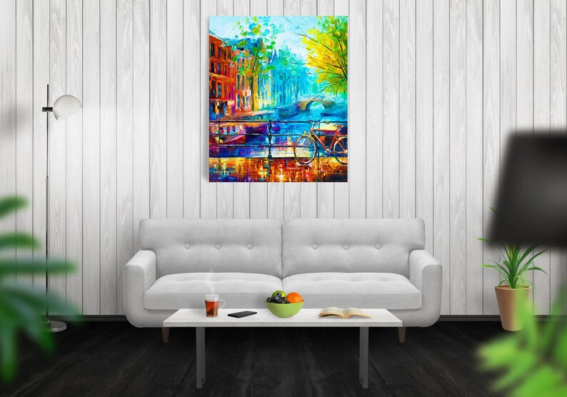 May include: An abstract painting of a European city scene with a bridge, buildings, and a bicycle. The artwork features vibrant colors, including blues, oranges, and yellows, creating a lively and artistic impression. The painting is displayed above a white sofa.