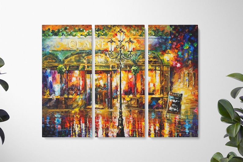May include: A triptych canvas print of a vibrant cityscape at night. The artwork depicts a cafe with the name "CLARE" above the entrance, a lamppost, and reflections on a wet street. The color palette includes warm tones of orange, yellow, and red.