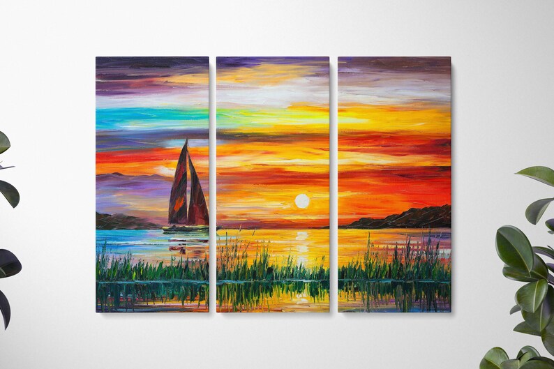 May include: A triptych canvas print depicting a sunset over water. The artwork features a sailboat with a brown sail, a bright yellow sun, and colorful sky with shades of orange, red, and blue. Green reeds are in the foreground.