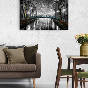 May include: A black and white painting of a bridge over a canal at night. The artwork depicts a European city scene with streetlights illuminating the water and buildings. The painting is displayed on a white wall.