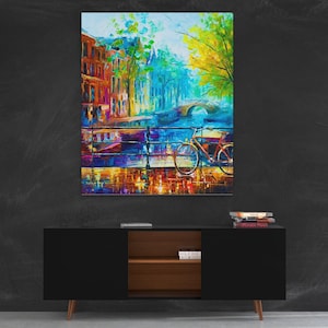 Pode incluir: Uma pintura a óleo vibrante retrata uma cena de canal com edifícios, uma ponte e uma bicicleta. A obra de arte apresenta uma paleta colorida de azuis, amarelos e vermelhos, criando uma sensação animada e impressionista. A pintura é exibida acima de um armário preto.