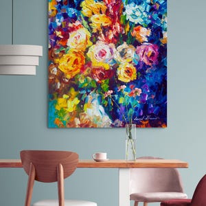 May include: An oil painting of a vibrant floral arrangement. The artwork features a bouquet of yellow, pink, and white roses against a deep blue background. The artist's signature is visible in the lower right corner. The painting is displayed on a wall above a wooden table.