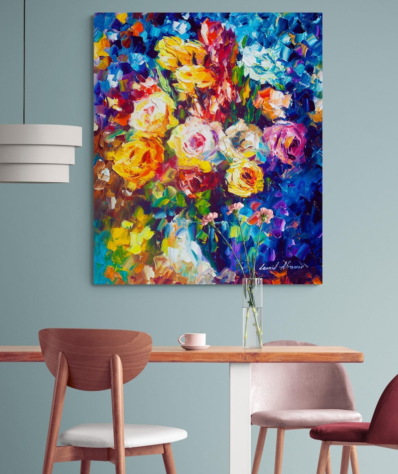 May include: An oil painting of a vibrant floral arrangement. The artwork features a bouquet of yellow, pink, and white roses against a deep blue background. The artist's signature is visible in the lower right corner. The painting is displayed on a wall above a wooden table.