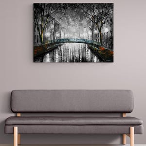 May include: A black and white painting of a canal scene with a bridge, trees, and buildings. The artwork is displayed above a gray sofa with wooden legs. The painting has a moody, atmospheric quality.