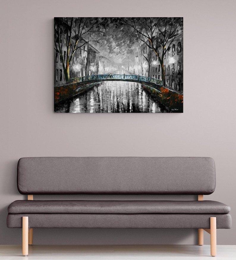 May include: A black and white painting of a canal scene with a bridge, trees, and buildings. The artwork is displayed above a gray sofa with wooden legs. The painting has a moody, atmospheric quality.