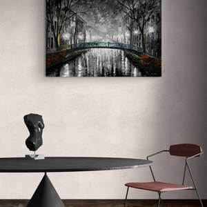 May include: A grayscale painting of a canal scene at night, featuring a bridge and buildings with lights reflecting in the water. The artwork is displayed above a black table with a sculpture and a chair.
