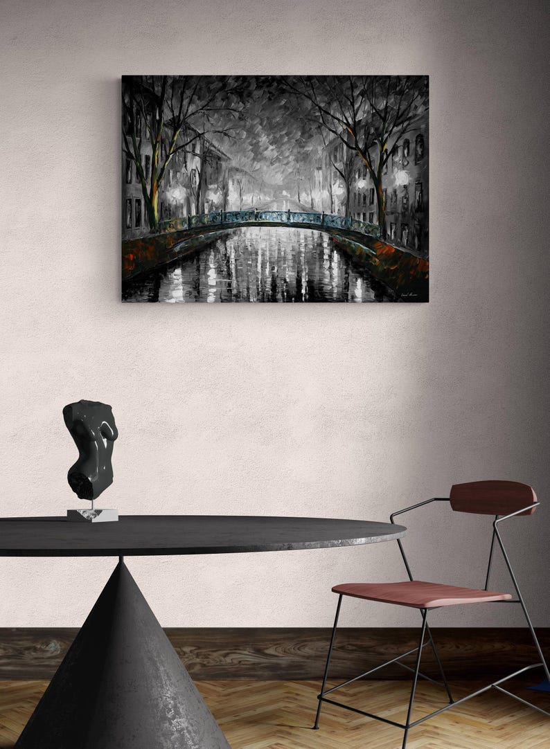 May include: A grayscale painting of a canal scene at night, featuring a bridge and buildings with lights reflecting in the water. The artwork is displayed above a black table with a sculpture and a chair.