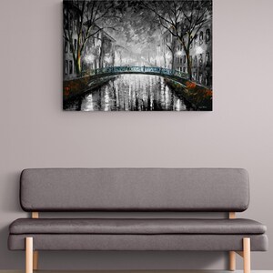 May include: A black and white oil painting of a canal scene with a bridge. The artwork depicts a nighttime view with streetlights illuminating the water and buildings. The painting is displayed above a gray sofa with wooden legs.