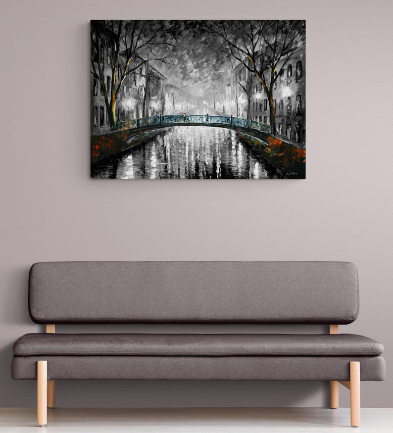 May include: A black and white oil painting of a canal scene with a bridge. The artwork depicts a nighttime view with streetlights illuminating the water and buildings. The painting is displayed above a gray sofa with wooden legs.