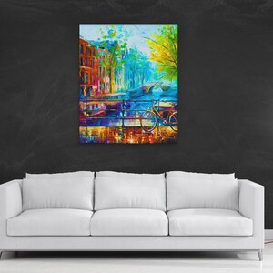 May include: A colorful abstract painting of a European cityscape, featuring buildings, a bridge, and a bicycle. The artwork uses vibrant blues, oranges, and greens, creating a lively and artistic scene. The painting is displayed above a white sofa.
