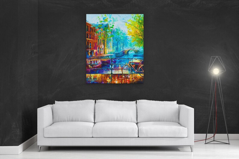 May include: A colorful abstract painting of a European cityscape, featuring buildings, a bridge, and a bicycle. The artwork uses vibrant blues, oranges, and greens, creating a lively and artistic scene. The painting is displayed above a white sofa.