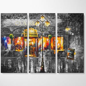 May include: A triptych canvas print of a nighttime street scene. The artwork is primarily in grayscale, with vibrant orange and yellow hues illuminating the shop windows. A decorative street lamp stands in front of the shop.