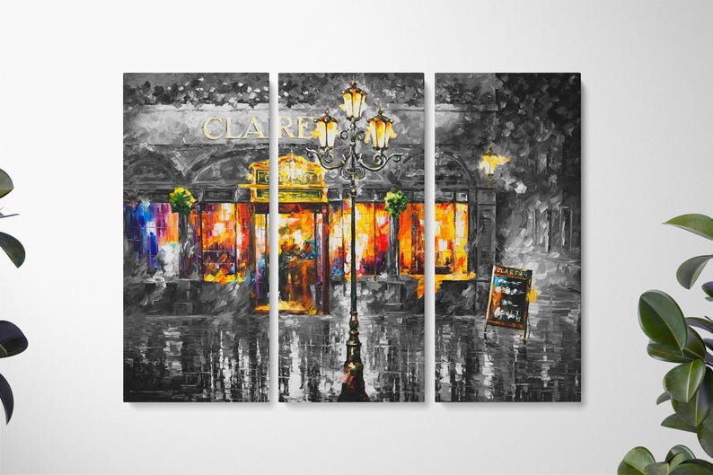May include: A triptych canvas print of a nighttime street scene. The artwork is primarily in grayscale, with vibrant orange and yellow hues illuminating the shop windows. A decorative street lamp stands in front of the shop.