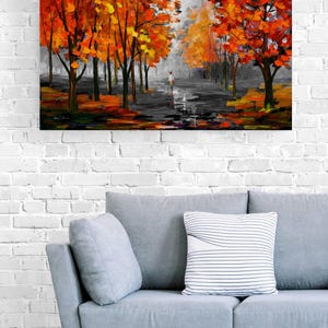 May include: An oil painting depicts a path lined with trees in autumn. The trees have vibrant orange and yellow leaves. A person walks down the path. The painting hangs above a gray sofa with pillows.
