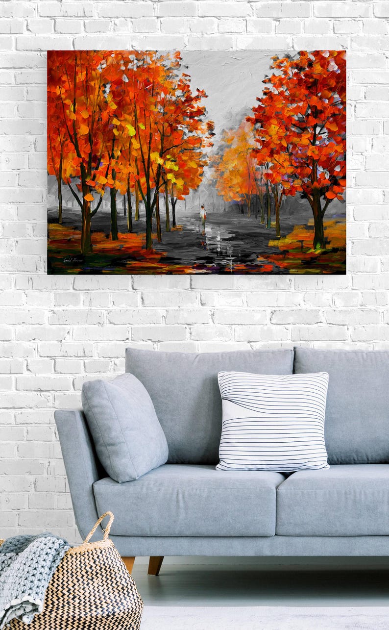 May include: An oil painting depicts a path lined with trees in autumn. The trees have vibrant orange and yellow leaves. A person walks down the path. The painting hangs above a gray sofa with pillows.