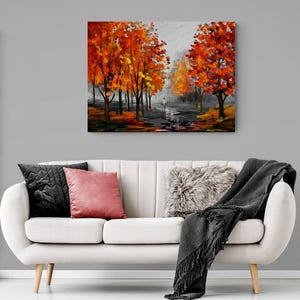 May include: An oil painting depicts a path lined with trees ablaze in autumn colors. The artwork features a person walking on the path, with a gray sky overhead. The painting hangs above a white sofa with pillows and a blanket.
