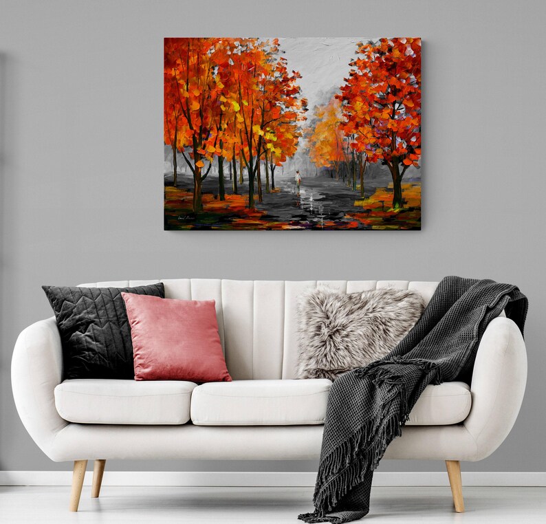 May include: An oil painting depicts a path lined with trees ablaze in autumn colors. The artwork features a person walking on the path, with a gray sky overhead. The painting hangs above a white sofa with pillows and a blanket.