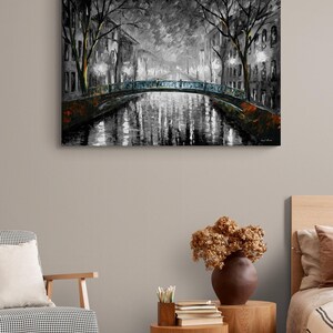 May include: A black and white painting of a canal scene at night. A bridge with blue accents spans the water, reflecting the lights of the buildings and trees along the canal. The artwork is displayed on a wall.