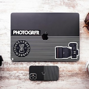 Pode incluir: Um laptop preto com um adesivo que diz "PHOTOGRFR" e um adesivo com uma x&iacute;cara de caf&eacute; e o texto "NEVER STOP BREWING". H&aacute; um adesivo de lente de c&acirc;mera preta no laptop. Um telefone preto com um design listrado est&aacute; na frente do laptop.