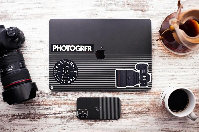Pode incluir: Um laptop preto com um adesivo que diz "PHOTOGRFR" e um adesivo com uma x&iacute;cara de caf&eacute; e o texto "NEVER STOP BREWING". H&aacute; um adesivo de lente de c&acirc;mera preta no laptop. Um telefone preto com um design listrado est&aacute; na frente do laptop.