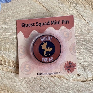 Könnte beinhalten: Eine runde, orange und schwarze Anstecknadel mit einem Drachen und dem Text "QUEST SQUAD" darauf. Die Anstecknadel befindet sich auf einer Karte mit dem Text "Quest Squad Mini Pin" und "Capturedbystories".