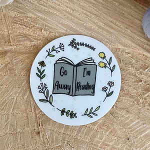 Puede incluir: Una pegatina redonda blanca con una ilustración de un libro y el texto "Go Away I'm Reading" rodeada de hojas verdes y flores amarillas.