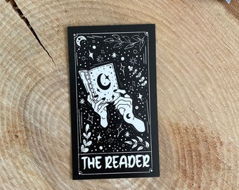 Puede incluir: Carta de tarot en blanco y negro con una mano sosteniendo un libro abierto con una luna creciente en la portada. La carta está rodeada de estrellas y el texto "THE READER" está impreso en la parte inferior.
