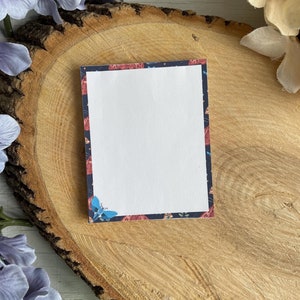 Puede incluir: Una tarjeta en blanco con un borde floral en tonos azules, rojos y blancos. La tarjeta está sobre una superficie de madera.