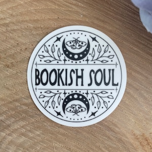 May include: A white round sticker with a black design. The design features a crescent moon with a moth, stars, and leaves. The text "BOOKISH SOUL" is written in a black font.