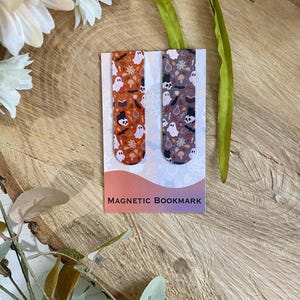 May include: Two magnetic bookmarks with Halloween patterns. One has an orange background with ghosts and bats. The other has a purple background with similar designs. The words "MAGNETIC BOOKMARK" are printed on the card.