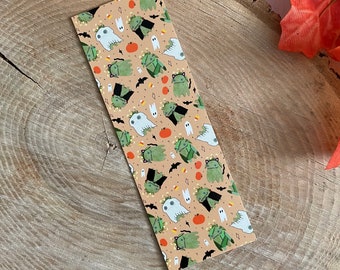 Könnte beinhalten: Ein Lesezeichen zum Thema Halloween mit einem sich wiederholenden Muster aus grünen Monstern, Geistern, Fledermäusen, Zuckermais und Kürbissen auf einem pfirsichfarbenen Hintergrund.