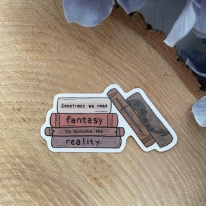 Op de afbeelding: Een sticker met een stapel boeken met een zwaard- en drakenontwerp. De tekst op de sticker luidt "Sometimes we need fantasy to survive the reality."