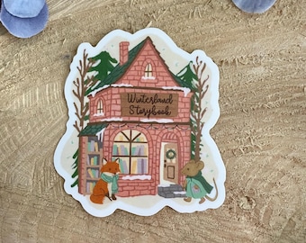 May include: A sticker featuring a whimsical illustration of a brick building with the text "Winterland Storybook." A fox wearing a scarf and a mouse in a cape stand near the entrance. The building has a green roof and is surrounded by trees.