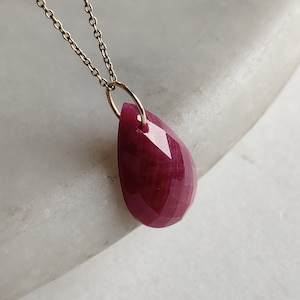 Puede incluir: Un collar con un colgante de rubí en forma de lágrima y una cadena de oro. El rubí es de color rojo intenso y tiene un corte facetado.