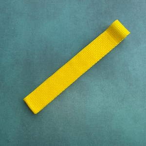 Op de afbeelding: Een felgele stoffen band, mogelijk een horlogeband of een vergelijkbaar accessoire, ligt diagonaal over een blauwgroen oppervlak. De band is gemaakt van geweven materiaal en het uiteinde is omgeslagen. De algehele esthetiek is eenvoudig en strak.