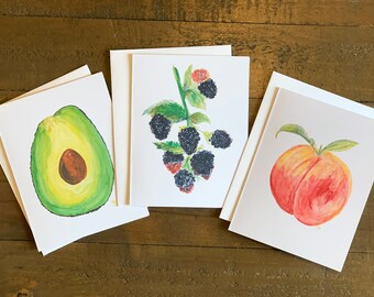 May include: Three watercolour greeting cards with fruit illustrations. One card shows a sliced avocado, another a branch of blackberries, and the third a ripe peach. Each card is paired with a blank envelope, set against a wooden background.
