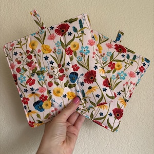 Puede incluir: Dos agarraderas con estampado floral sobre fondo blanco. La tela presenta un colorido patrón de flores rojas, amarillas, azules y rosas, junto con hojas verdes y pequeñas bayas rojas. Cada agarradera tiene un pequeño lazo de tela para colgar.