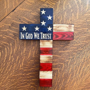 K&ouml;nnte beinhalten: Ein Holzkreuz, das mit der amerikanischen Flagge verziert ist. Das Kreuz besteht aus einzelnen Holzbrettern, die in Rot, Wei&szlig; und Blau gestrichen sind. Die Worte "In God We Trust" sind auf das Kreuz gedruckt.