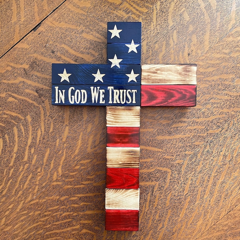 K&ouml;nnte beinhalten: Ein Holzkreuz, das mit der amerikanischen Flagge verziert ist. Das Kreuz besteht aus einzelnen Holzbrettern, die in Rot, Wei&szlig; und Blau gestrichen sind. Die Worte "In God We Trust" sind auf das Kreuz gedruckt.