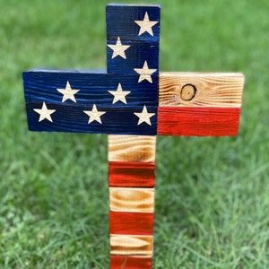 K&ouml;nnte beinhalten: Holzkreuz, das in den Farben der amerikanischen Flagge bemalt ist. Das Kreuz ist aus wiederverwendetem Holz gefertigt und hat einen rustikalen Look. Die Oberseite des Kreuzes ist blau mit wei&szlig;en Sternen bemalt, die Mitte ist rot und die Unterseite wei&szlig;.