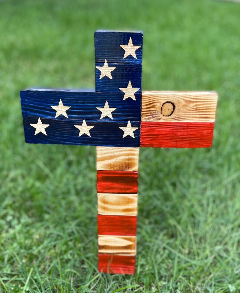K&ouml;nnte beinhalten: Holzkreuz, das in den Farben der amerikanischen Flagge bemalt ist. Das Kreuz ist aus wiederverwendetem Holz gefertigt und hat einen rustikalen Look. Die Oberseite des Kreuzes ist blau mit wei&szlig;en Sternen bemalt, die Mitte ist rot und die Unterseite wei&szlig;.