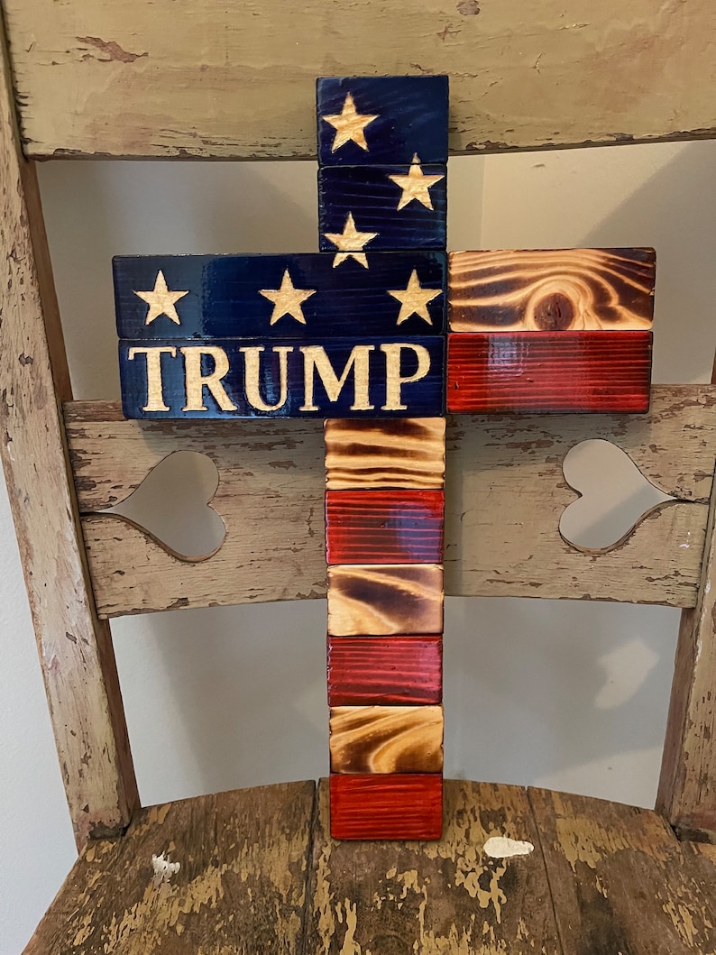 K&ouml;nnte beinhalten: Holzkreuz mit amerikanischer Flaggen-Optik. Das Kreuz besteht aus einzelnen Holzbrettern, die in Rot, Wei&szlig; und Blau gestrichen sind. Das Wort "TRUMP" ist in wei&szlig;en Buchstaben auf dem blauen Teil des Kreuzes geschrieben.