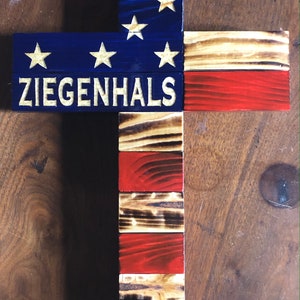 K&ouml;nnte beinhalten: Ein Holzkreuz mit amerikanischer Flaggen-Optik. Das Kreuz ist blau und rot mit wei&szlig;en Sternen und Streifen bemalt. Die Worte "ZIEGENHALS" sind in Wei&szlig; auf das Kreuz geschrieben.