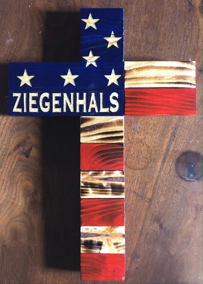 K&ouml;nnte beinhalten: Ein Holzkreuz mit amerikanischer Flaggen-Optik. Das Kreuz ist blau und rot mit wei&szlig;en Sternen und Streifen bemalt. Die Worte "ZIEGENHALS" sind in Wei&szlig; auf das Kreuz geschrieben.