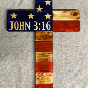 K&ouml;nnte beinhalten: Holzkreuz mit amerikanischer Flaggen-Optik. Das Kreuz ist in Rot, Wei&szlig; und Blau lackiert mit blauem Hintergrund und wei&szlig;en Sternen. Die Worte "JOHN 3:16" sind in Gold auf das Kreuz geschrieben.