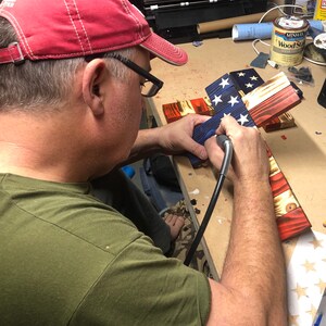 K&ouml;nnte beinhalten: Eine Person arbeitet an einem Holzkreuz mit der amerikanischen Flagge. Die Flagge ist in Rot, Wei&szlig; und Blau bemalt. Die Person verwendet ein Werkzeug, um die Sterne auf der Flagge zu detaillieren.