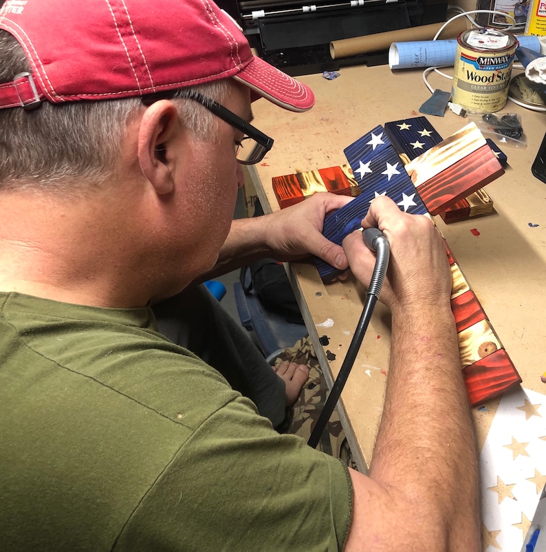 K&ouml;nnte beinhalten: Eine Person arbeitet an einem Holzkreuz mit der amerikanischen Flagge. Die Flagge ist in Rot, Wei&szlig; und Blau bemalt. Die Person verwendet ein Werkzeug, um die Sterne auf der Flagge zu detaillieren.