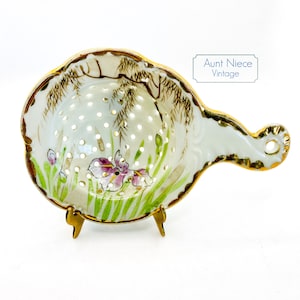 May include: A white porcelain tea strainer with a gold rim and handle. The strainer is decorated with a floral design and a gold-painted willow tree. The strainer is on a gold metal stand.